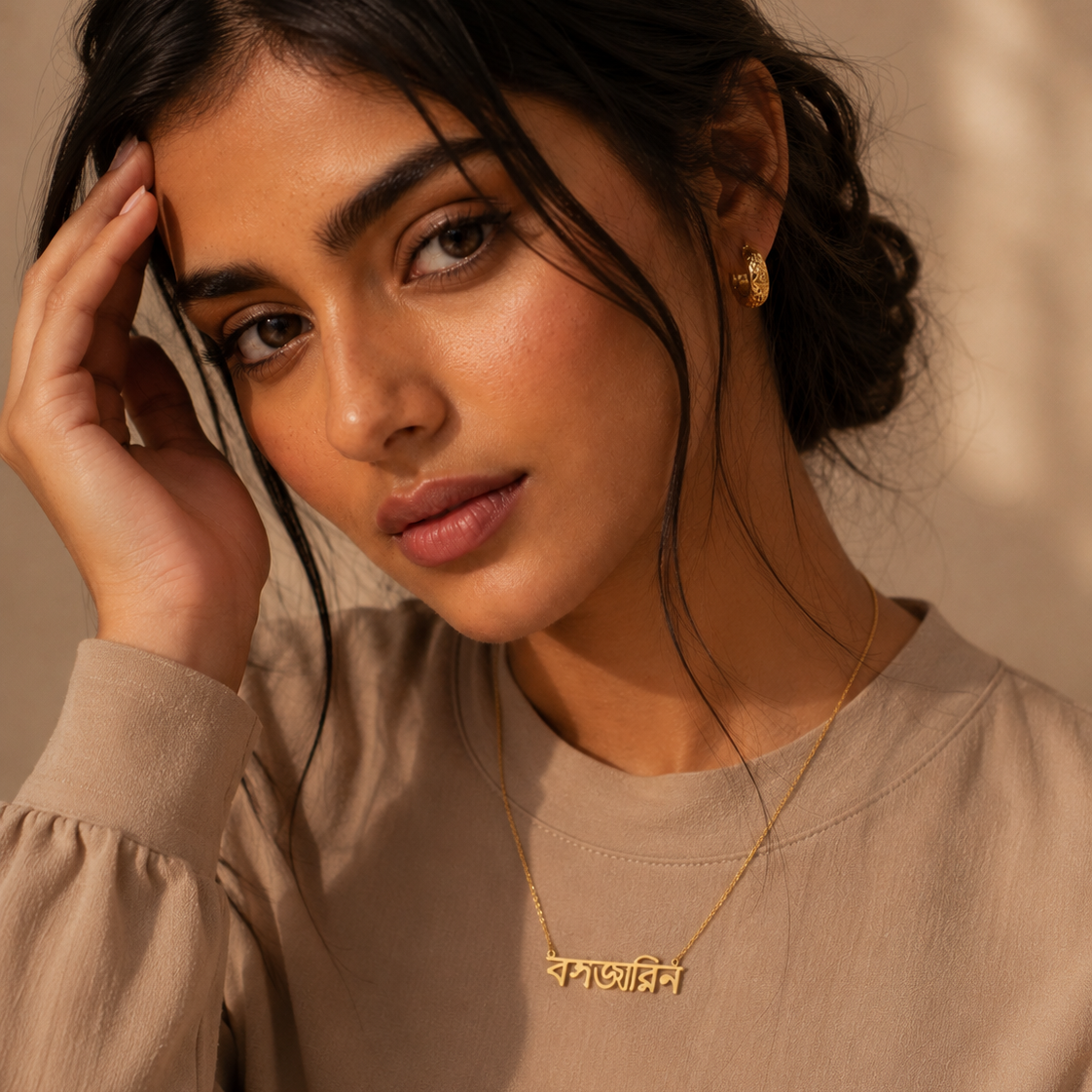 A woman wearing a delicate gold necklace with a Bangla script nameplate and a small gold stud earring, paired with a beige long-sleeve top against a warm beige background.