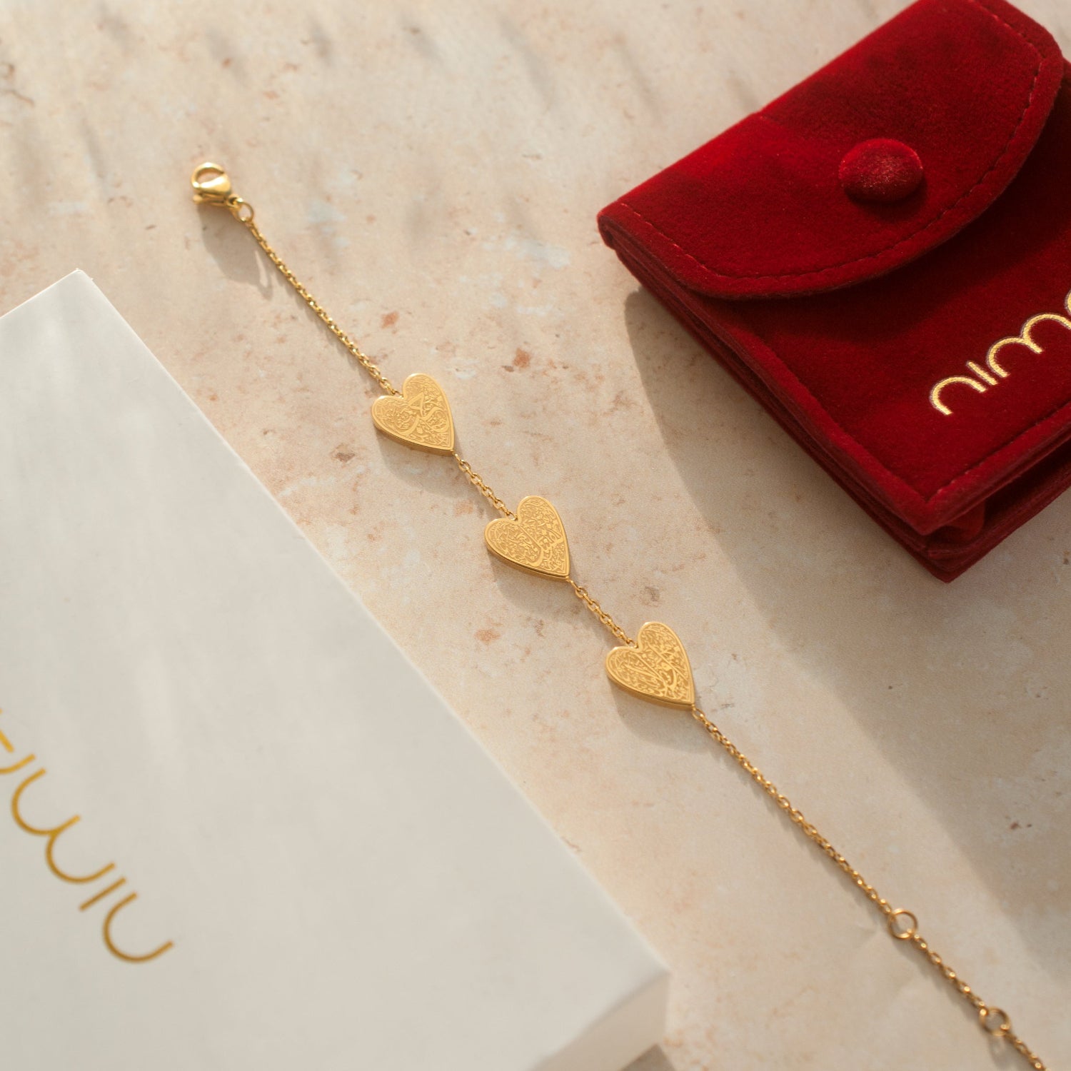 Gold heart-shaped bracelet on a textured surface with a red pouch and white box.