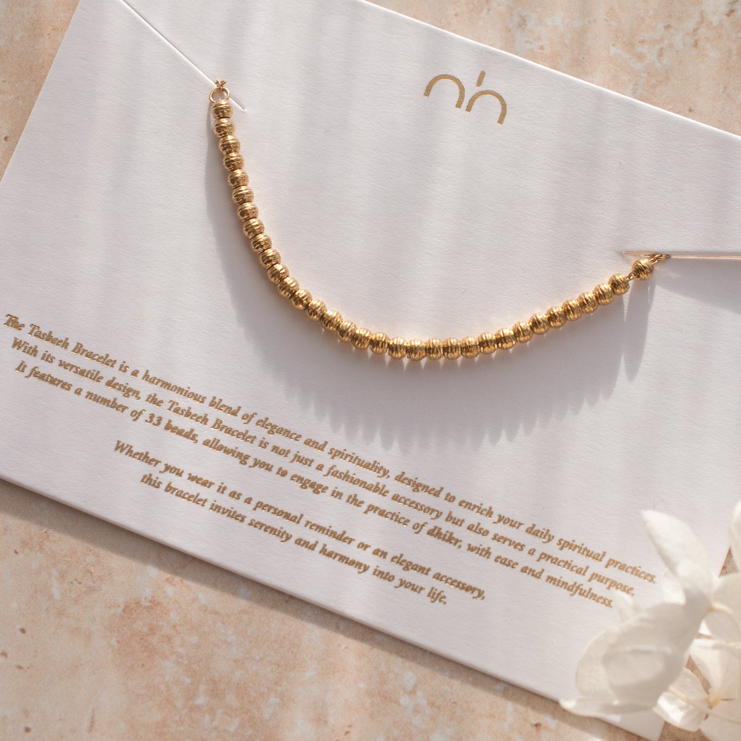Gold tasbih bracelet on a white card with text and nimah logo, on a beige surface