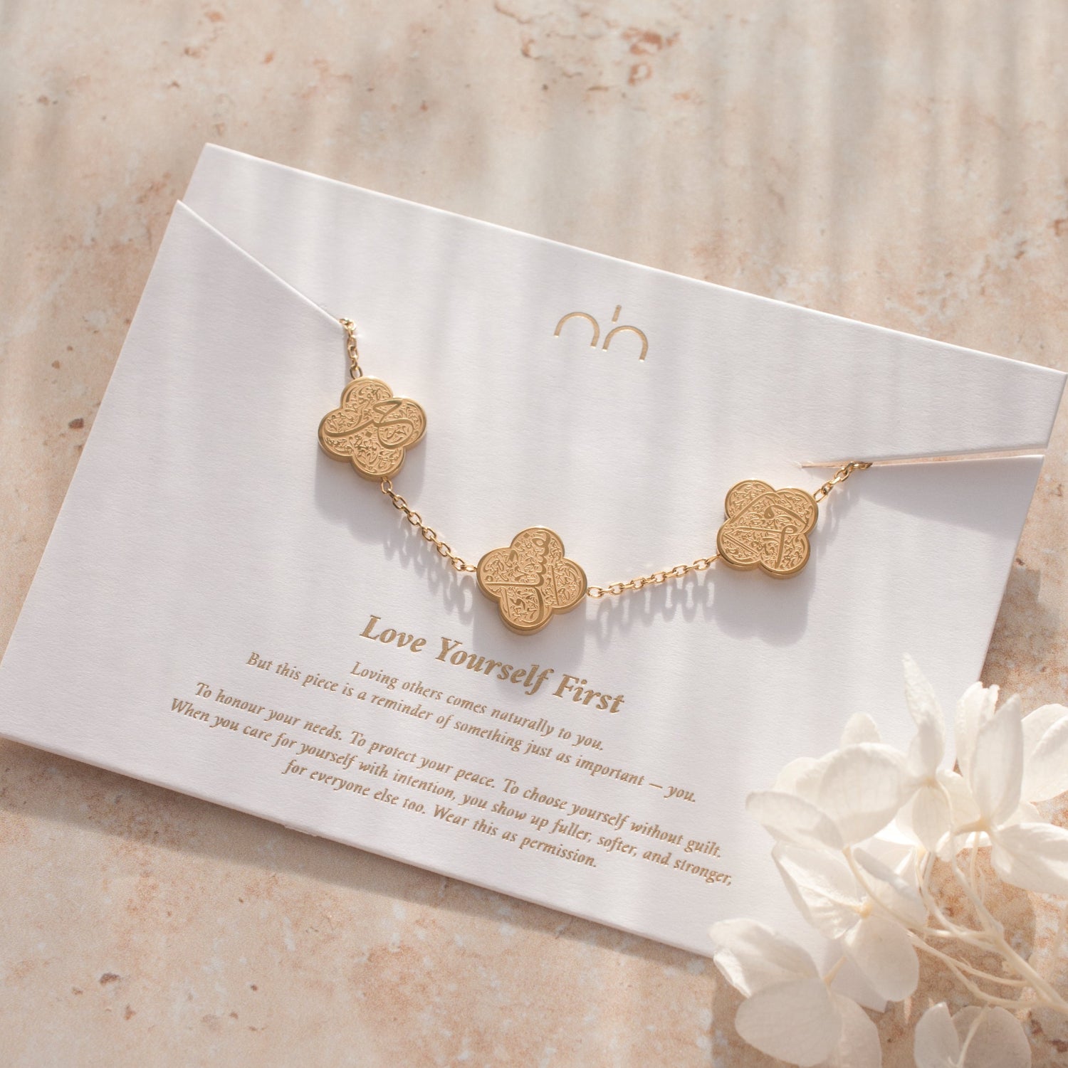 Gold necklace with clover-shaped pendants on a card with text, placed on a textured surface with flowers.
