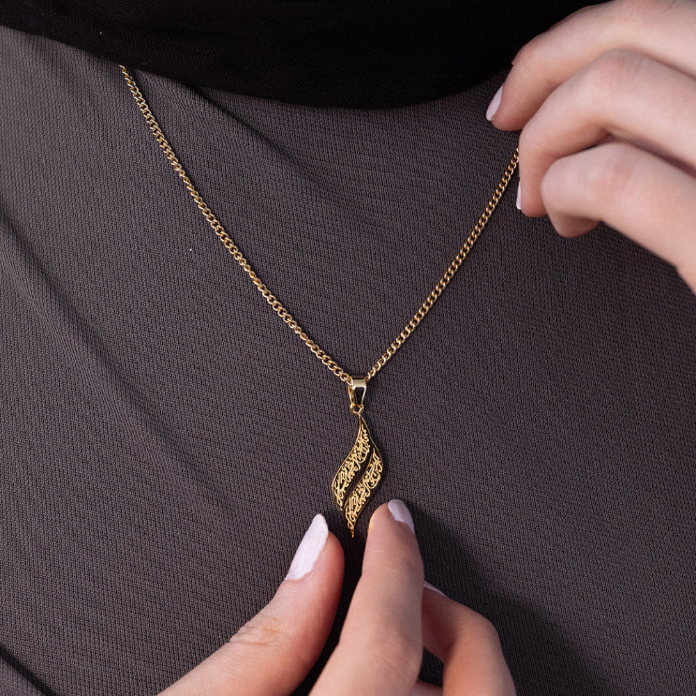A close-up of a person wearing a gold chain necklace with a textured, wavy diamond-shaped pendant, styled with a matching gold ring against a dark gray textured background.