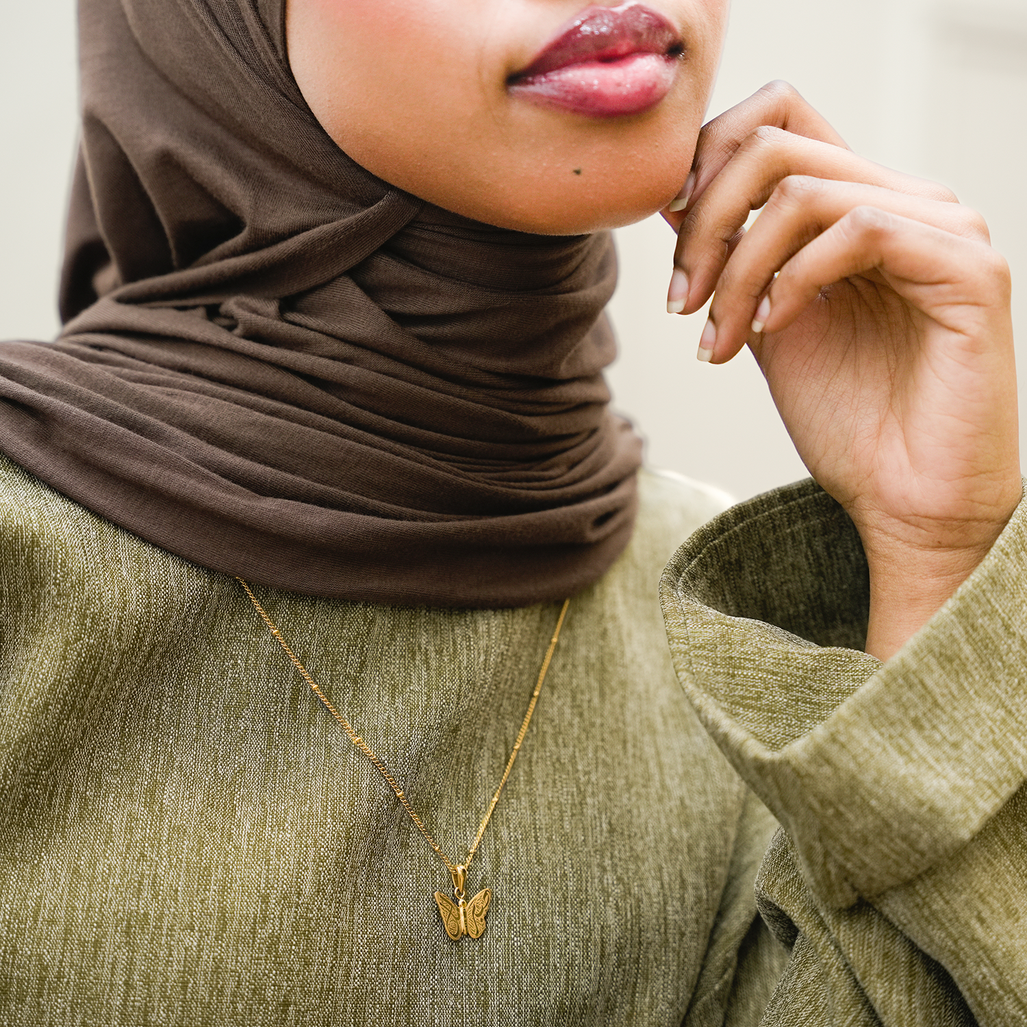 Person wearing a brown hijab and green sweater with a gold necklace featuring a butterfly pendant.