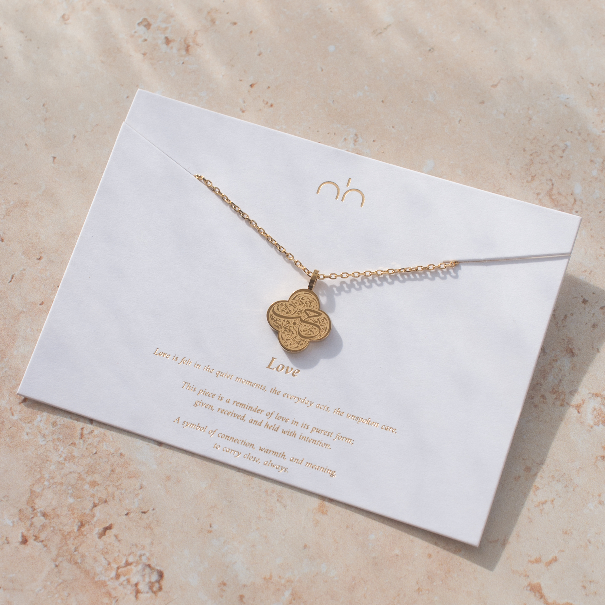 Gold necklace with a heart-shaped pendant on a white card with gold text, placed on a beige surface.