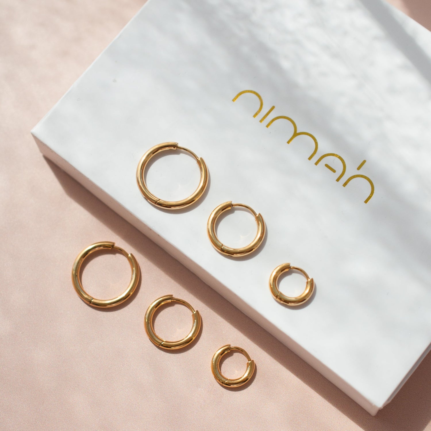 Gold hoop earrings on a white box with 'nimah' branding on a pink background