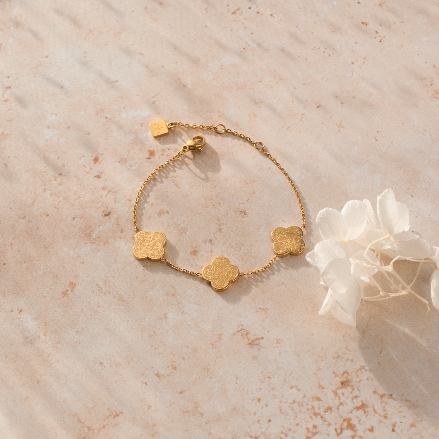 Nimah Gold bracelet with clover-shaped arabic writing charms on a beige surface with a white flower.