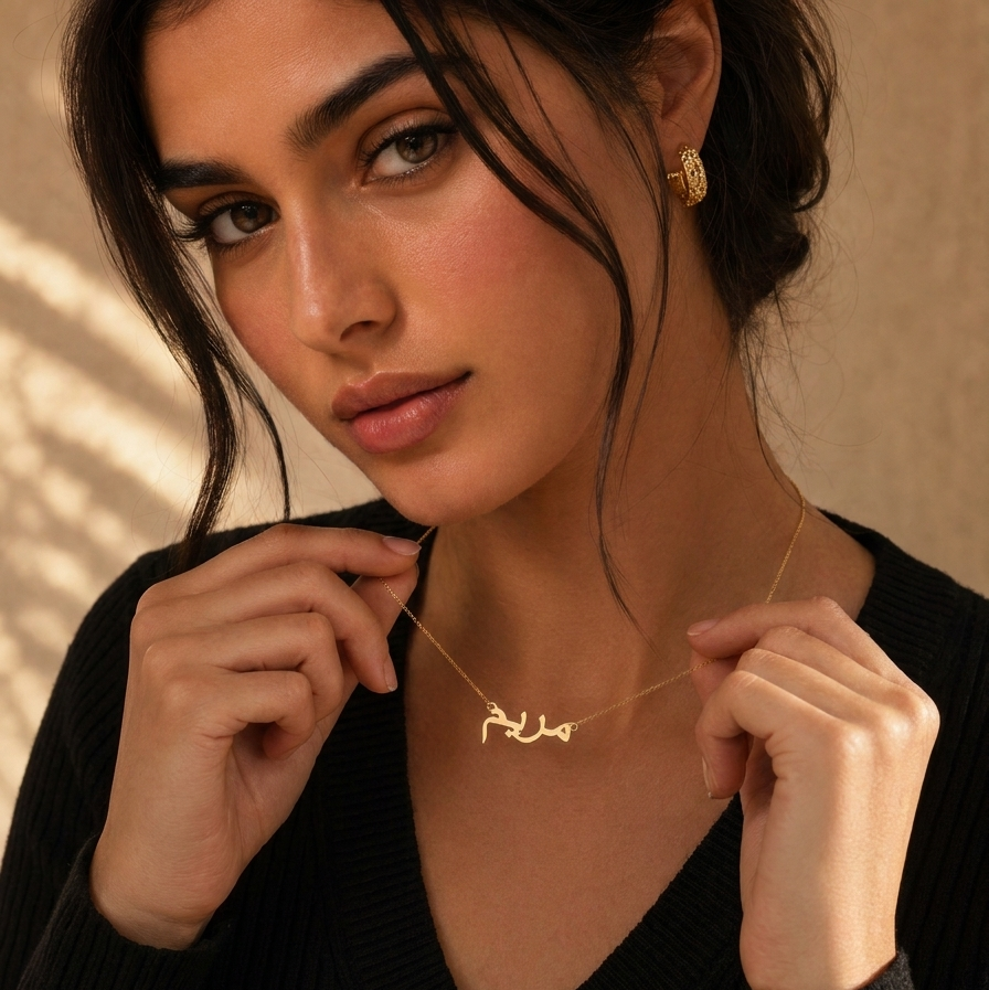 A close-up lifestyle shot of a woman wearing a delicate gold necklace with Arabic calligraphy script, holding it near her collarbone with her hands. She is wearing a black ribbed V-neck sweater and a small gold earring, set against a warm beige background with soft leaf shadows.