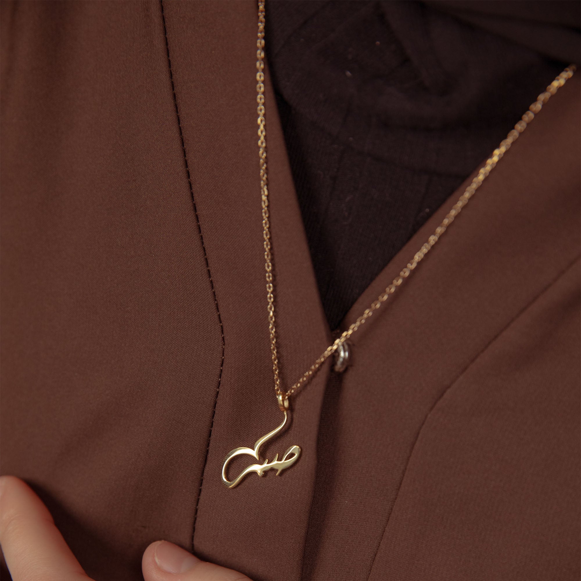 Gold "Patience" Arabic calligraphy necklace worn over a brown abaya